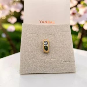 Anillo dorado Yanbal con piedra central sobre exhibidor, fondo de flores rosadas.