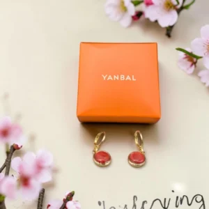 Aretes dorados Yanbal con dije coral, junto a su caja original y flores decorativas.