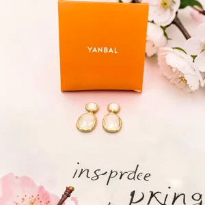 Aretes dorados Yanbal con detalle blanco, junto a su caja original y flores decorativas.