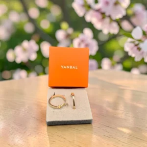 Candongas doradas Yanbal sobre cojín gris, junto a caja naranja de la marca, con flores rosadas de fondo.