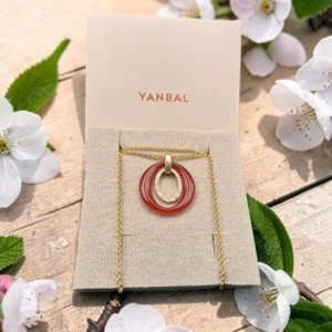 Collar Yanbal con dije ovalado rojo y cadena dorada sobre empaque original, entre flores blancas.