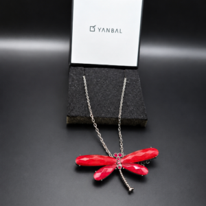 Collar Yanbal con dije de libélula roja sobre caja y base negra de exhibición.