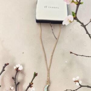 Collar dorado Yanbal con dije de luna azul, presentado junto a su caja original y flores.