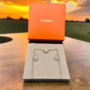 Collar y topitos Yanbal con dijes azules sobre exhibidor, caja Yanbal de fondo con atardecer.