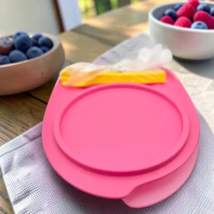 Contenedor redondo rosado con tapa sobre mesa de madera, junto a frutas