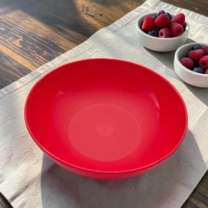 Bol plástico redondo color rojo sobre mesa de madera, con moras y frambuesas al fondo