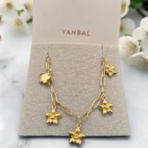Pulsera dorada con dijes de flores exhibida en tarjeta Yanbal sobre fondo con flores blancas.
