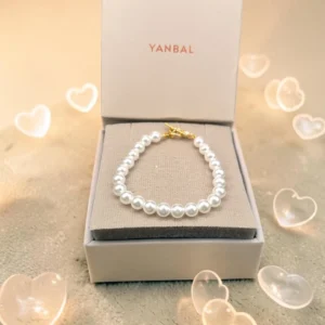 Pulsera de perlas blancas con cierre dorado dentro de caja Yanbal, decoración de corazones alrededor.