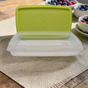 Contenedor rectangular Tupperware transparente con tapa verde sobre mesa de madera, con frutas de fondo.