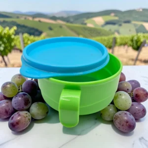 Taza alimentadora verde con tapa azul y asa, rodeada de uvas