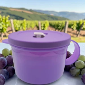 Taza hermética redonda color lila con asa y tapa, rodeada de uvas.